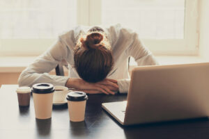 A woman with her head on her desk, behind a laptop and several coffee cups