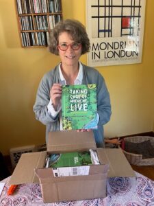 Woman opening a box of books and holding up a copy of a book titled Taking Care of Where We Live.