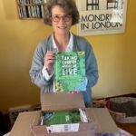 Woman Opening A Box Of Books And Holding Up A Copy Of A Book Titled Taking Care Of Where We Live.