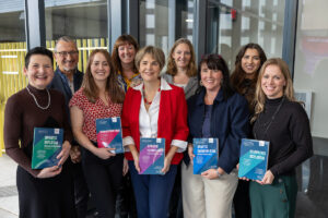 A group of nine smiling people holding books, with WCEA partner Ruth Wilson wearing a red jacket and white blouse. 